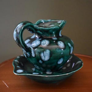 May include: A small, green ceramic pitcher with a matching saucer. The pitcher has a handle and a ruffled rim. The surface is glazed with a dark green color and white polka dots. The set is likely vintage.