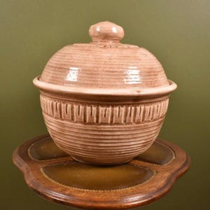 May include: A light brown ceramic lidded bowl with a textured, ribbed design. The bowl has a rounded shape with a decorative band near the base. The lid features a small knob and a matching ribbed pattern. The bowl is sitting on a wooden tray.