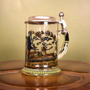 May include: A clear glass beer stein with a hinged metal lid and handle. The stein features a painted scene with a figure, deer, and floral elements. The base has a yellow and black decorative border. The text on the stein reads "...mir mein verla..."