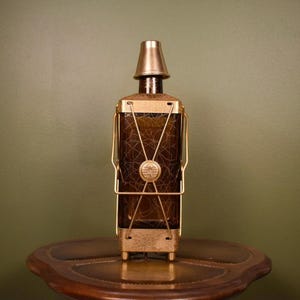 May include: A decorative brown glass bottle with a gold-colored metal cage and a matching conical stopper. The bottle is rectangular with a textured pattern. The metal cage has a circular emblem and a base with small feet.