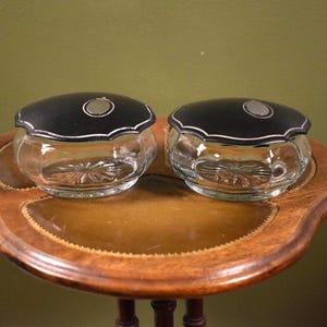 May include: Two matching glass vanity jars with clear, faceted bases and scalloped edges. Each jar features a smooth, matte black lid with a central circular silver-toned inlay. The glass bodies have a starburst pattern on the bottom.