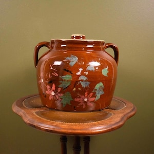 May include: A brown ceramic crock with a lid and two handles, decorated with floral and leaf designs in pink, blue, and green. The crock sits on a small, round wooden table. A small knob is on top of the lid.