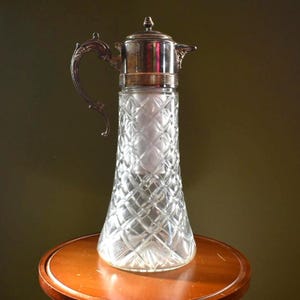 May include: A tall, clear glass pitcher with a diamond-cut pattern. It has a silver-colored metal top, handle, and spout. The pitcher is displayed on a round, wooden table, against a dark green background. The pitcher is likely used for serving beverages.