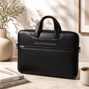 Può includere: Una borsa porta computer nera di forma rettangolare con due manici superiori, una tasca frontale con cerniera e il nome ALEXANDER WILLIAMS impresso in lettere dorate. La borsa presenta ganci in metallo argentato per la tracolla.