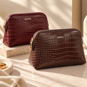 May include: Two dome-shaped cosmetic pouches with a crocodile-embossed texture. The back pouch is deep red with the name OLIVIA in gold lettering, and the front pouch is dark brown with the name EMILY in gold lettering, both featuring gold zippers.