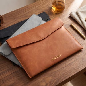 May include: A stack of three leather document holders in brown, gray, and black. The brown holder is on top, with the name "CAITLYN" embossed in gold. The holders are on a wooden surface.