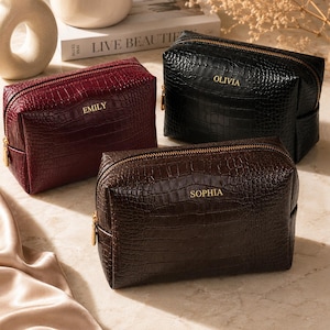May include: Three rectangular cosmetic bags with a crocodile-embossed texture in burgundy, black, and dark brown. Each bag features a gold-tone zipper and is personalized with gold lettering: EMILY on the burgundy, OLIVIA on the black, and SOPHIA on the brown.