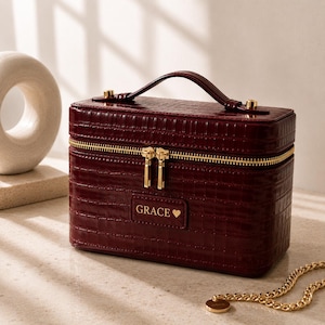 May include: A rectangular, deep burgundy vanity case with a crocodile-embossed texture. It features a gold-toned zip closure, a top handle, and a rectangular patch on the front with the text "GRACE" followed by a small heart symbol.