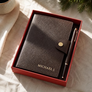 May include: A dark brown textured notebook with a gold snap closure and the name MICHAEL J. embossed in gold lettering on the cover, accompanied by a matching dark brown pen with gold accents, all presented inside a rectangular red gift box.
