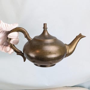 May include: A metallic, dark-toned teapot with a rounded body, a curved handle, and a tapered spout. The teapot features a central lid finial and a smooth, polished surface with visible signs of age and patina throughout the metal finish.