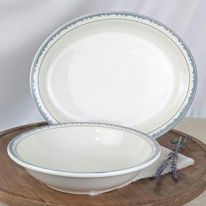 May include: Two-piece white ceramic serving dish set. The oval platter and round bowl have a light blue scalloped border with a thin dark blue line. The set is displayed on a wooden tray.