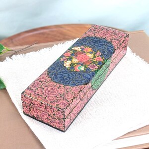 May include: A rectangular box with a hinged lid featuring a dense floral pattern in shades of pink, blue, and green. The surface is covered in intricate, hand-painted flower motifs that repeat across the top and sides of the elongated container.