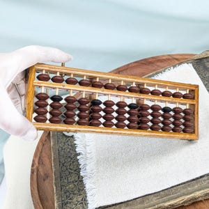 May include: A wooden abacus featuring a rectangular frame with vertical rods. The device contains rows of brown beads, with a few black beads interspersed among them. The beads are oval-shaped and arranged in a grid pattern within the wooden structure.