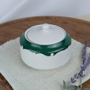 May include: A white ceramic sugar bowl with a lid, accented with a glossy green band around the top. The bowl sits on a light gray linen napkin, with a wooden surface in the background. A sprig of lavender is in the foreground.