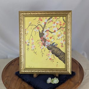 May include: Framed painting of a flowering tree branch with red, white, and pink blossoms, and two birds perched on the branch. The artwork is set against a yellow background and framed in a gold-colored frame.
