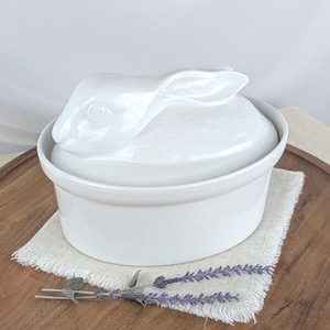 May include: White ceramic casserole dish with a rabbit-shaped lid. The dish is round with a smooth, glossy finish. The lid features a detailed rabbit head. The dish sits on a beige linen napkin and a wooden tray. A sprig of lavender is placed near the dish.