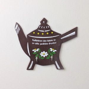 May include: A brown wooden teapot-shaped key holder with white accents and the text "Kaffetåren den bästa är av alla jordiska drycker". It features a floral design and two hooks for keys.