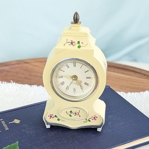 May include: A cream-colored mantel clock with a rounded top and a pointed finial. The clock face features black Roman numerals and gold-toned hands. The exterior is decorated with small hand-painted pink flowers and green leaves on the front.