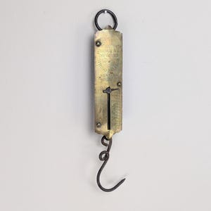 May include: A vintage brass spring balance scale featuring a dark metal hook at the bottom and a circular ring at the top. The face is engraved with the text CHATILLON'S IMPROVED SPRING BALANCE NEW YORK and includes a vertical sliding indicator.
