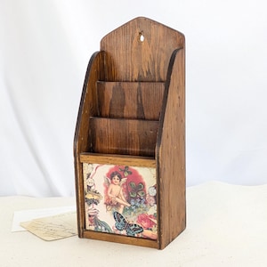 May include: Vintage wooden mail organizer with three compartments. The front features a decorative panel with a cherub, butterflies, and floral elements. The wood has a warm, natural finish, and the organizer has a triangular top with a small hole for hanging.