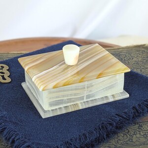 May include: A rectangular onyx box with a lid and a small cylindrical knob. The lid features a striped pattern in shades of beige and brown. The box is set on a dark blue cloth.