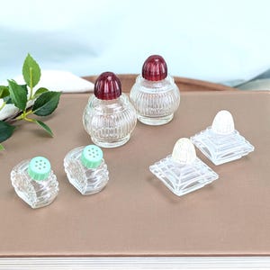 May include: A collection of six glass shakers featuring two round clear glass shakers with ribbed sides and dark red caps, two round clear glass shakers with ribbed sides and light green caps, and two square clear glass shakers with white caps.