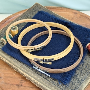 May include: Three circular embroidery hoops of varying sizes and wood tones, each featuring a metal tension screw mechanism. The hoops are stacked on top of a piece of dark blue fabric with frayed edges, which rests on a vintage book with a decorative cover.