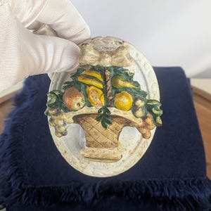 May include: An oval-shaped, off-white wall plaque featuring a basket overflowing with colorful fruit. The fruit includes yellow bananas, peaches, and grapes. The basket is brown, and the plaque has a decorative bow at the top.