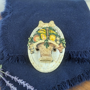 May include: An oval-shaped, off-white wall plaque featuring a basket overflowing with colorful fruits. The basket is brown, and the fruits include yellow lemons, red apples, and green grapes. A decorative bow sits atop the plaque.