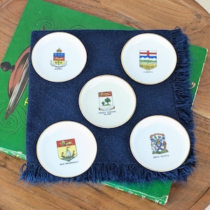 May include: Five small, white plates with gold rims, each featuring a different provincial crest. The plates are arranged on a dark blue fringed cloth. The crests represent Quebec, Alberta, Prince Edward Island, New Brunswick, and Nova Scotia.
