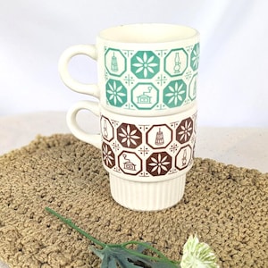 May include: Two stacked ceramic mugs with a repeating geometric pattern of octagons containing floral motifs, coffee grinders, and oil lamps. The top mug features teal graphics, while the bottom mug displays brown graphics on a cream-colored base.