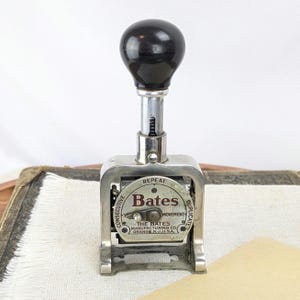 May include: Vintage Bates Manufacturing Co. stamp with a black handle and silver metal frame. The stamp features the text "Bates" and "The Bates Manufacturing Co. Orange, N.J. U.S.A." on a circular dial. The stamp is designed for consecutive, repeat, and duplicate imprints.