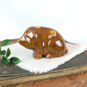 May include: A brown ceramic pig figurine with glossy finish and gold-colored spots. The pig is sitting on a white cloth, placed on top of a vintage book. The figurine has a rounded body and a detailed snout.