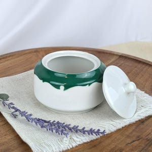 May include: A white ceramic sugar bowl with a dark green rim and lid. The lid is off to the side. The bowl is sitting on a white linen napkin on a wooden tray. A sprig of lavender is in the foreground.