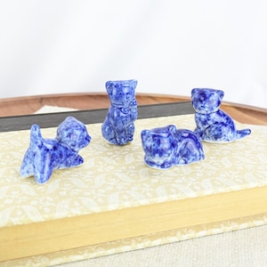 May include: Four small blue and white cat figurines in various poses. The cats are made of a speckled material and are arranged on a patterned surface. The figurines are a decorative item.
