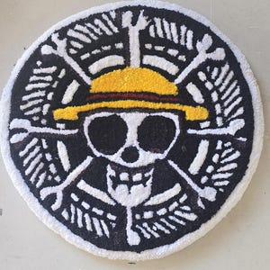 May include: Circular rug with a skull and crossbones design, wearing a yellow straw hat with a red band. The rug has a black background with white and yellow details. The rug's diameter is approximately 75 cm.
