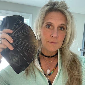 Può includere: Una donna con lunghi capelli biondi tiene un mazzo di carte da tarocchi nere con disegni bianchi. Indossa una camicia blu chiaro e una collana a strati con un ciondolo di pietra bianca.