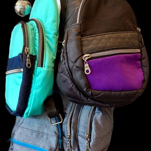 May include: A collection of four sling backpacks featuring a bright turquoise bag with dark blue accents, a black quilted bag with a purple front pocket, and two grey textured bags with blue and silver zippers, all equipped with metal hardware.