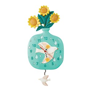 May include: A turquoise blue vase with a clock face and a white bird with yellow accents. The vase has a bouquet of yellow sunflowers on top. The clock face has black numbers and a black pendulum with a white bird.
