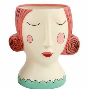 May include: A white ceramic planter shaped like a woman's face with closed eyes, pink cheeks, and red lips. The planter has pink hair with a swirl design and a teal green base.