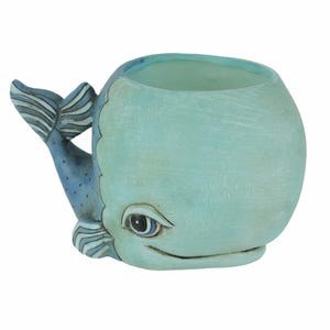 May include: A light blue whale planter with a blue and white tail fin. The whale has a painted eye and a small mouth.
