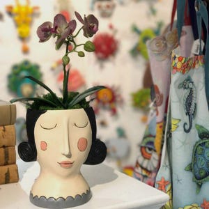May include: A white ceramic planter shaped like a woman's face with closed eyes and pink cheeks. The planter has a grey rim and is holding a small orchid plant with purple flowers.
