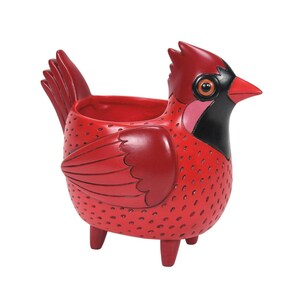 May include: A red cardinal-shaped planter with a detailed design. The bird has a black face, a pink patch, and a red beak. The body is covered in small, textured dots. The planter has a tail and wing details.