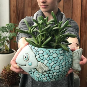 May include: A blue ceramic fish-shaped plant pot, filled with green succulents. The pot features a scale pattern and a smiling face. A decorative item for the home or garden, with a unique design.