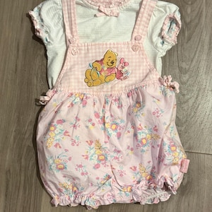 May include: A white short-sleeved shirt with a small bow at the neckline paired with pink and white gingham overalls. The overalls feature an embroidered Winnie the Pooh and Piglet graphic and a floral print on the lower portion with ruffled hems.