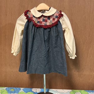 May include: A vintage-style baby dress with a cream-colored collar and sleeves. The bodice is navy blue with white polka dots, and the yoke features a patchwork design with red trim. The dress is displayed on a silver stand.