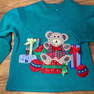 May include: Teal green sweatshirt with an appliqued teddy bear wearing a plaid vest and bow tie, sitting in a red box. The design includes gift boxes and ornaments with bows, perfect for the holiday season.