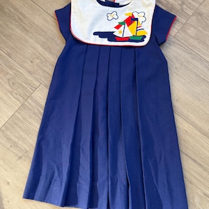 May include: A navy blue dress with a white collar featuring a sailboat design. The collar is trimmed with red piping, and the dress has short sleeves with red trim. The skirt is pleated, and the dress appears to be made of a soft fabric.
