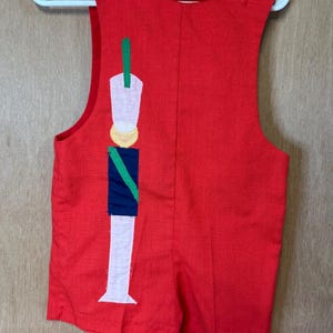 Puede incluir: Romper rojo con un diseño de soldado. El romper sin mangas presenta un diseño de soldado blanco, azul, verde y amarillo en la parte delantera. Dos botones blancos aseguran cada tirante. La prenda está colgada de una percha blanca.