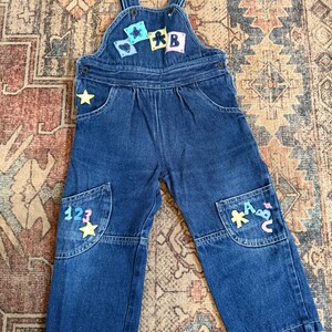 Op de afbeelding: Donkerblauwe denim tuinbroek voor peuters, met kleurrijke patches met letters, cijfers en sterren. De slab heeft patches met de letters A, B en een ster. De zakken hebben de cijfers 1, 2, 3 en de letters A, B, C.